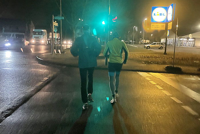 Zwei Läufer überqueren eine Kreuzung bei grüner Ampel im morgendlichen Dunkel