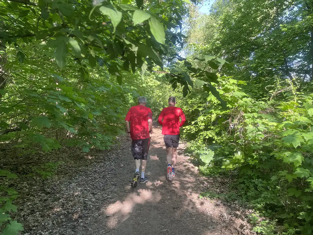 Zwei Läufer mit roten Laufshirts auf einem schmalen Weg, von allen Seiten umgeben von grünen Sträuchern und Bäumen